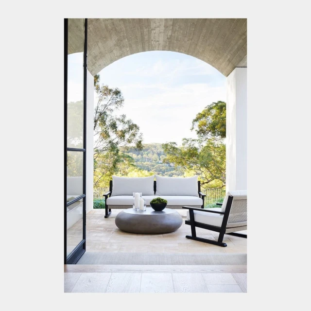 Outdoor seating area featuring a neutral-toned sofa, armchair, and round concrete coffee table with green plants, set against a hillside view.