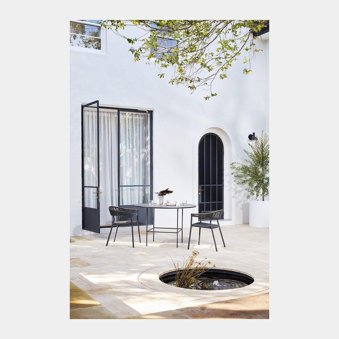Outdoor dining setting with a black table and two chairs on a stone patio, bathed in natural light with a minimalist architectural backdrop.