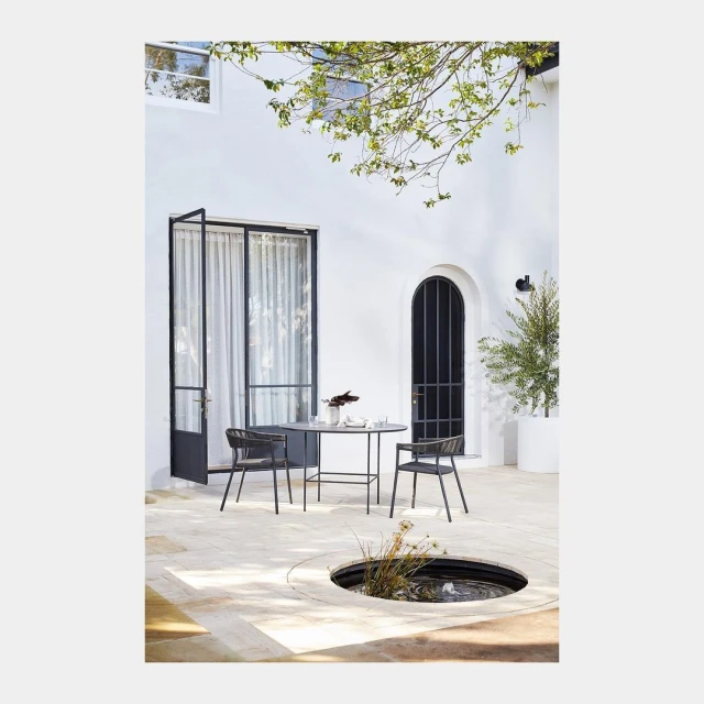 Outdoor dining setting with a black table and two chairs on a stone patio, bathed in natural light with a minimalist architectural backdrop.