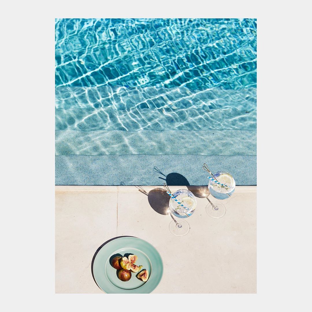 Aerial view of a turquoise swimming pool, drinks in wine glasses with straws, and figs on a plate, presenting a refreshing summer scene.