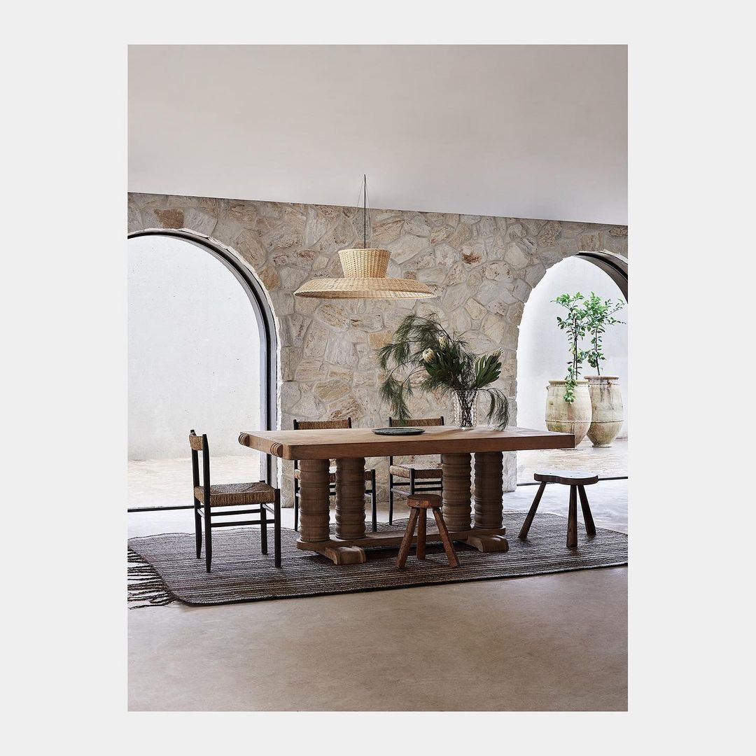 Interior view of a dining room with a long wooden table, chairs, a woven pendant light, stone walls, and a neutral color palette.
