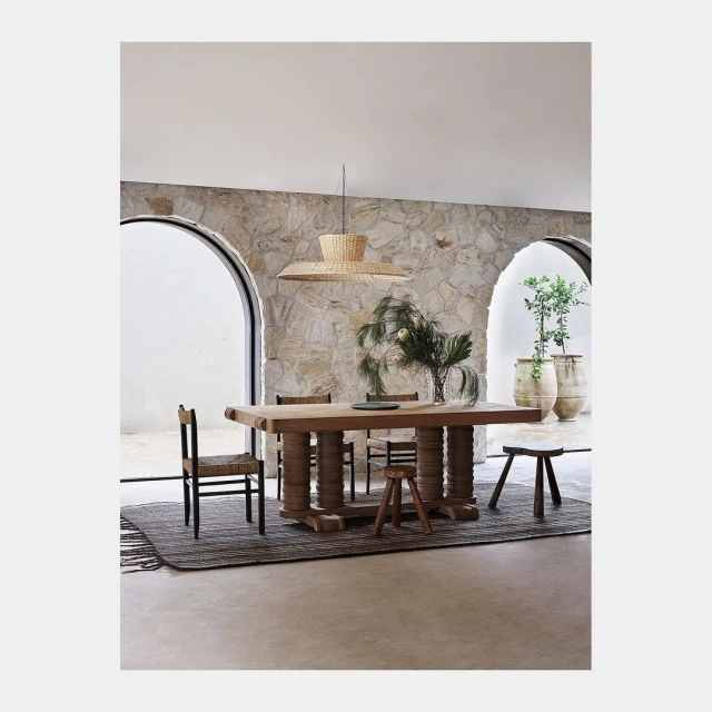 Interior view of a dining room with a long wooden table, chairs, a woven pendant light, stone walls, and a neutral color palette.