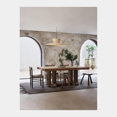 Interior view of a dining room with a long wooden table, chairs, a woven pendant light, stone walls, and a neutral color palette.