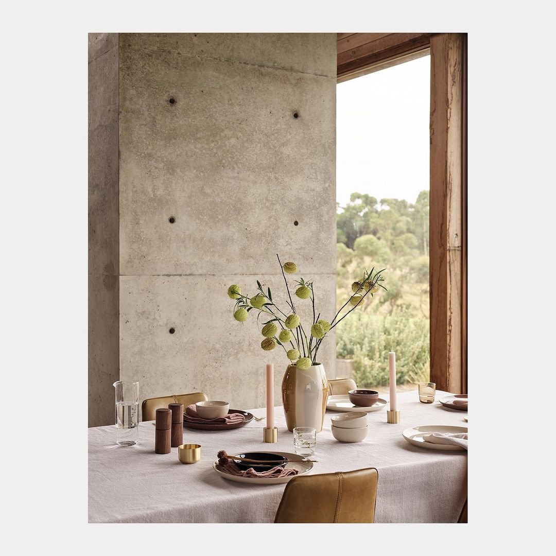 Still life shot of a table setting with beige plates, a vase with green leaves, glasses, candles, and brown chairs in a neutral room.