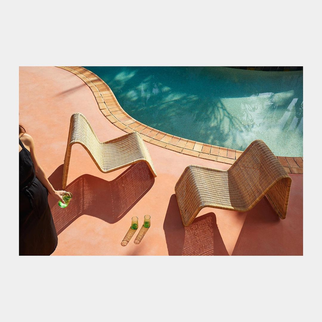 Two woven lounge chairs and clear glasses sit on a terracotta patio beside a turquoise swimming pool with dappled sunlit shadows.