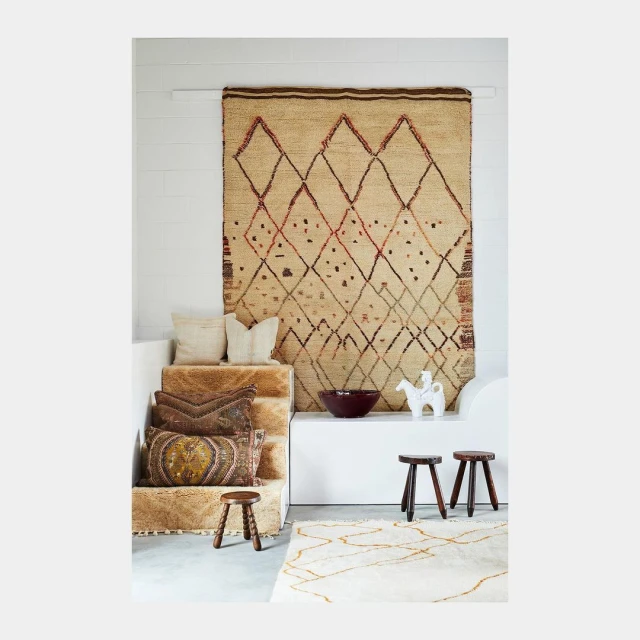 Cozy interior scene featuring an ornate, neutral-toned rug hanging on a white wall, complemented by cushions, stools, and decorative objects.