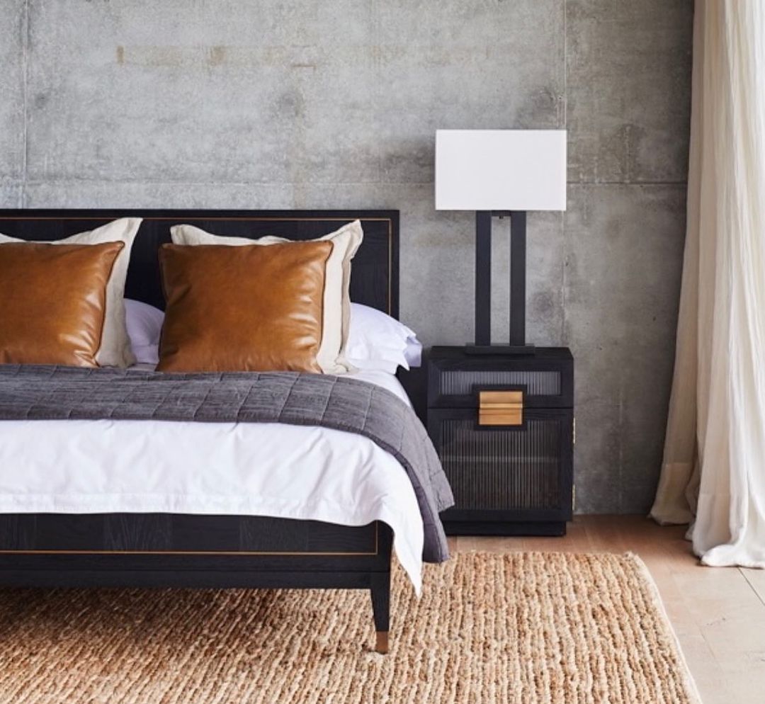 Bedroom interior featuring a bed with leather pillows, a nightstand with a lamp, a textured rug, and a neutral color scheme.