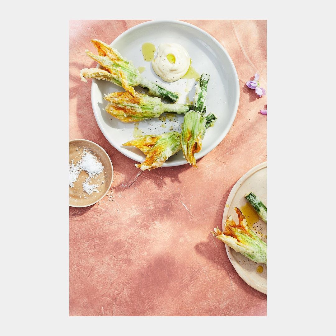 Overhead shot of two plates with fried zucchini flowers and white sauce against a pink textured backdrop, bathed in soft natural light.