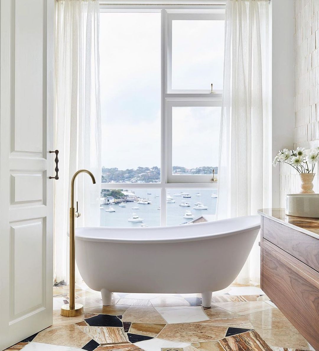 Bright bathroom interior showcasing a modern bathtub, brass fixtures, and large window overlooking a harbor with boats.