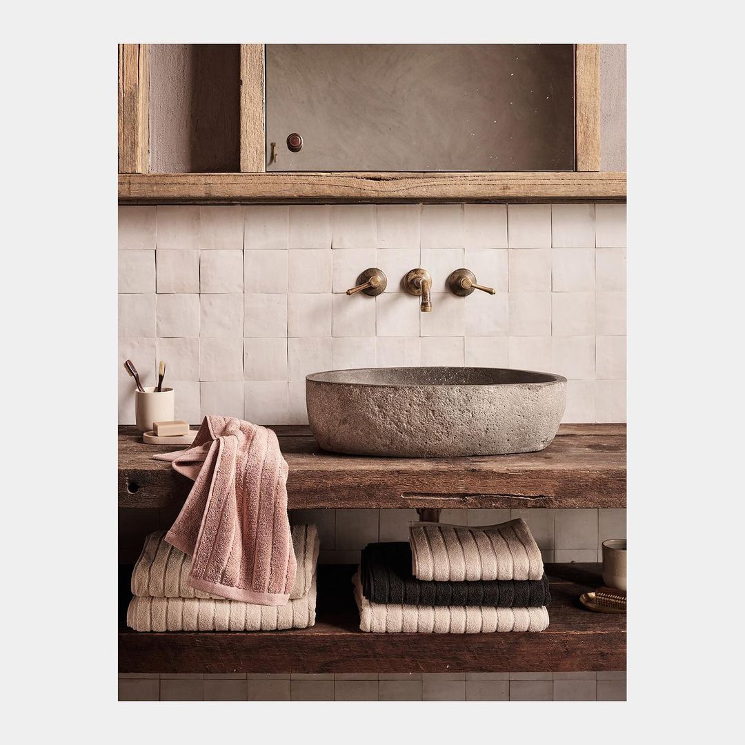 Bathroom interior featuring stone sink, rustic wood counter, neutral square tile wall, bronze faucet, and stacked, neatly folded towels.
