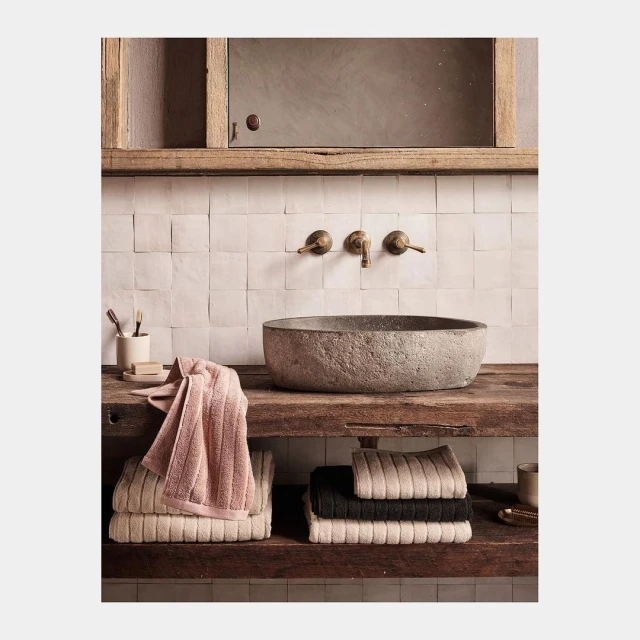 Bathroom interior featuring stone sink, rustic wood counter, neutral square tile wall, bronze faucet, and stacked, neatly folded towels.