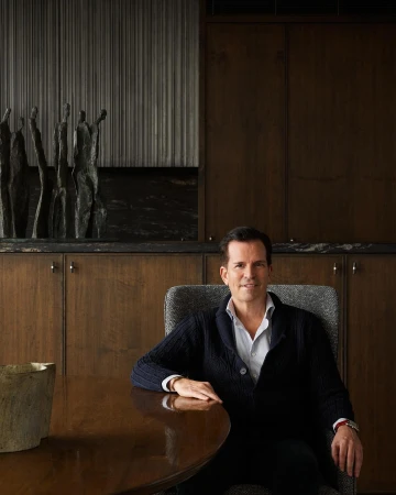 Man seated at a wooden table in an interior space with dark wood paneling and sculptural art.