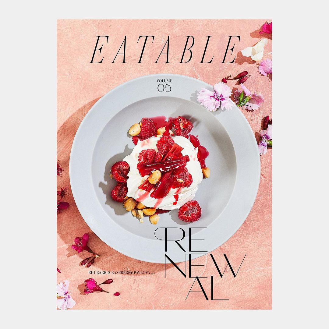 Overhead shot of a food magazine cover featuring a dessert topped with raspberries, styled with delicate flowers on a textured surface.
