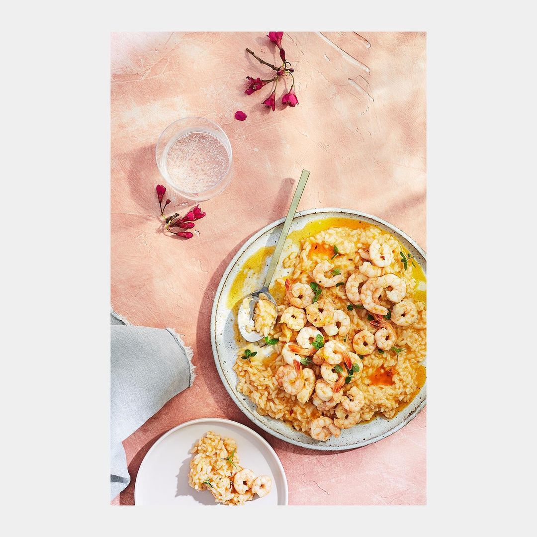 Overhead shot of shrimp risotto on plates, complemented by a glass of water and a delicate floral sprig on a textured pink surface.