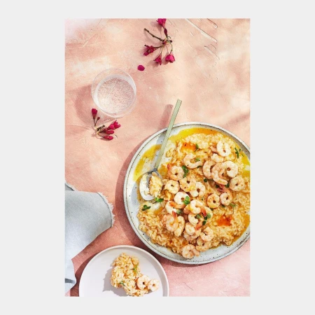 Overhead shot of shrimp risotto on plates, complemented by a glass of water and a delicate floral sprig on a textured pink surface.