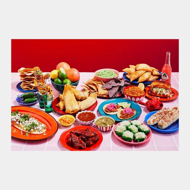 Overhead shot of a lavish Mexican food spread featuring tacos, tamales, chips, salsa, elote, and various side dishes on a pink tablecloth.