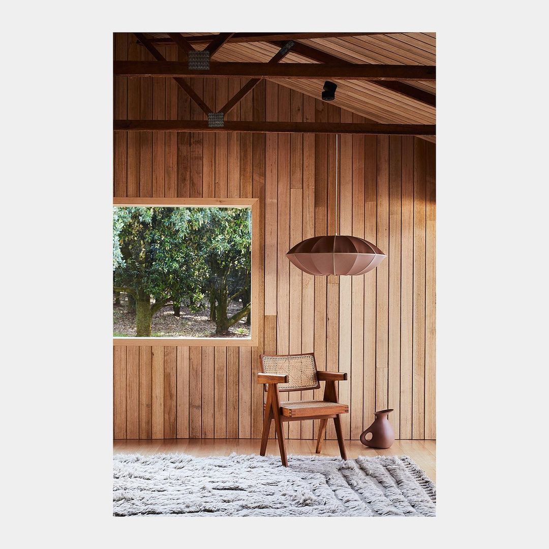 Interior view with light brown wooden walls and beamed ceiling, a window view of trees, chair, lamp, rug, and small vase.