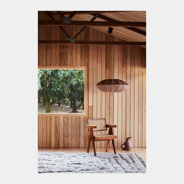 Interior view with light brown wooden walls and beamed ceiling, a window view of trees, chair, lamp, rug, and small vase.