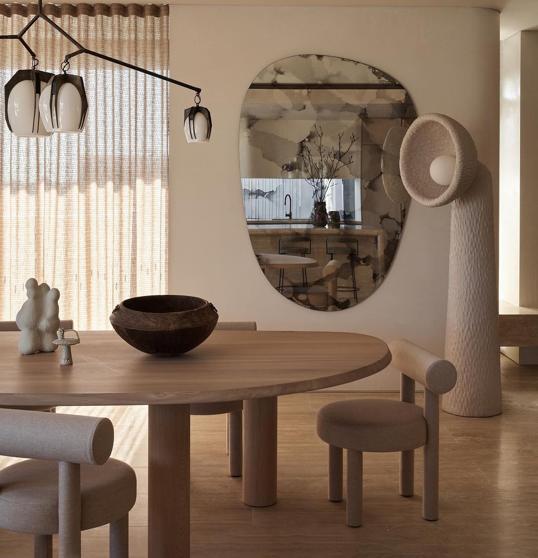 Modern dining room featuring a round wooden table with chairs, a sculptural lamp, and a unique mirror reflecting the kitchen interior.