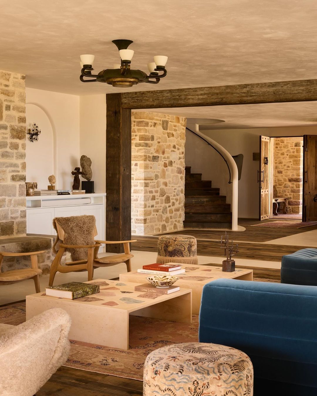 Living room interior showcasing neutral stone walls, wood accents, a blue sofa, patterned ottomans, and a light wood coffee table.