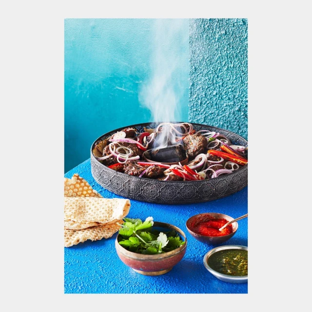 Close-up of smoking kabobs with red onions and peppers served in decorative metal plate with flatbread and three bowls of sauce.