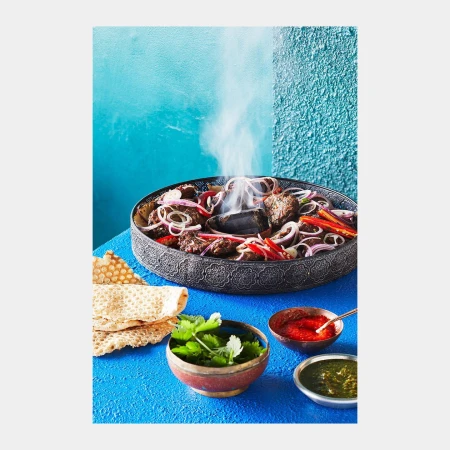 Close-up of smoking kabobs with red onions and peppers served in decorative metal plate with flatbread and three bowls of sauce.