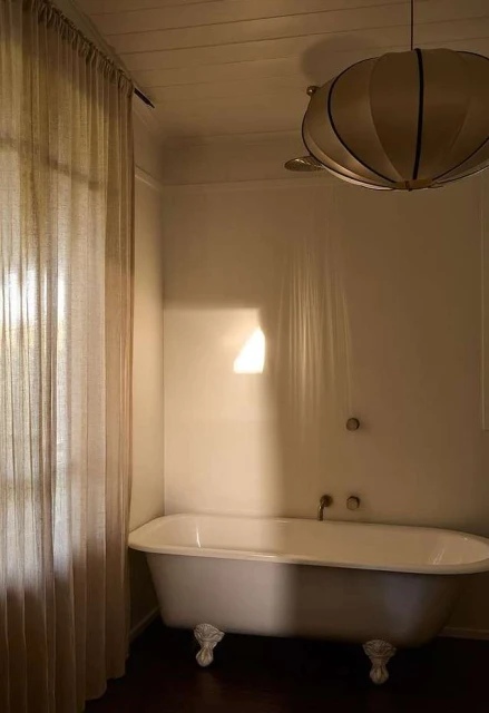 Clawfoot bathtub with white curtains and a unique ceiling light fixture in an interior bathroom with natural light and neutral tones.