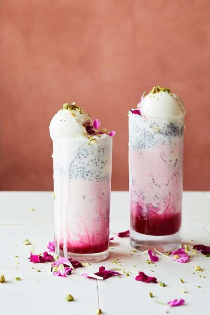Two glasses of Falooda, a South Asian layered dessert drink, topped with ice cream, nuts, and flower petals on a white table.