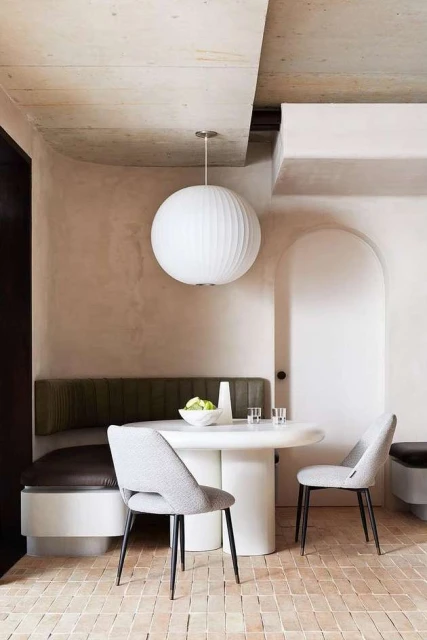 Indoor dining area featuring a white round table, two grey upholstered chairs, and a built-in bench, illuminated by a round pendant light.
