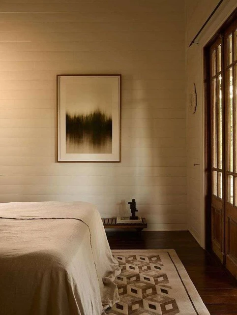 Beige bedroom features a bed with linen bedding, a geometric patterned rug, and framed artwork in a home interior space.
