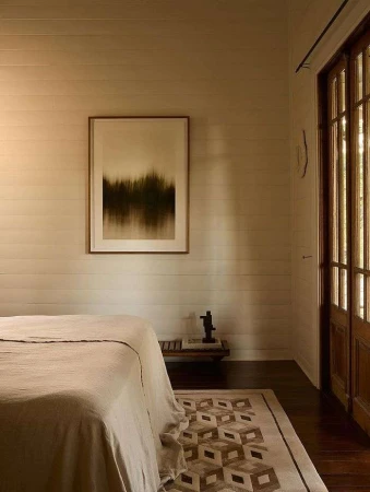 Beige bedroom features a bed with linen bedding, a geometric patterned rug, and framed artwork in a home interior space.