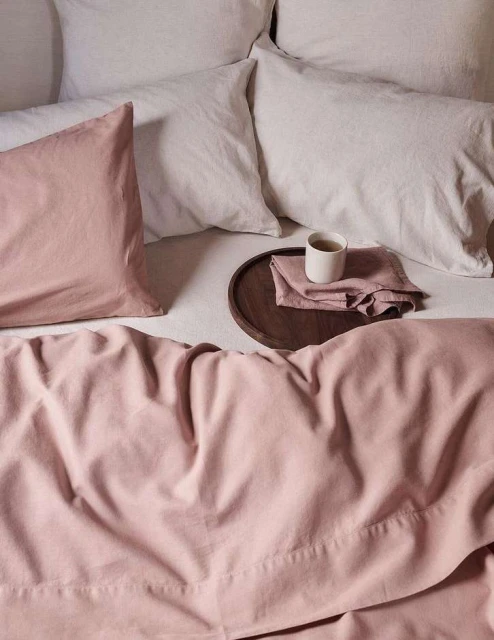 An inviting scene of a bed with light pink and white linens, pillows, and a wooden tray with a cup, styled for a cozy and serene bedroom setting.