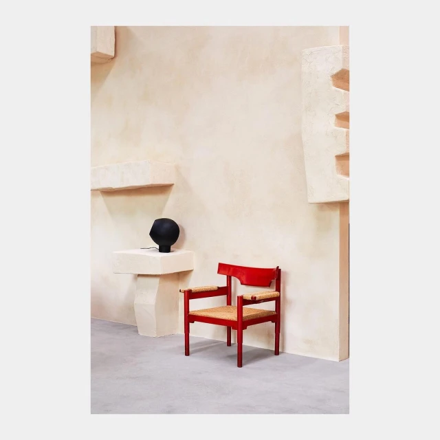 Red wooden chair with woven seat next to a black matte vase on a white plinth, against a minimalist beige wall.