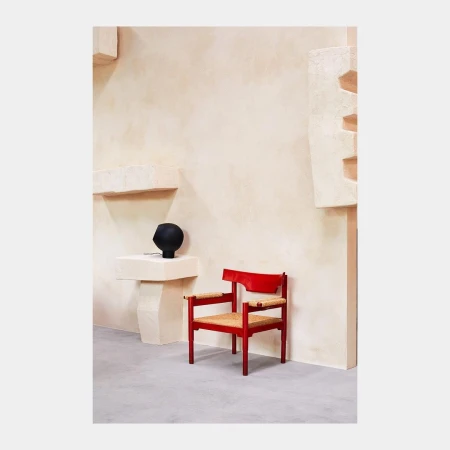 Red wooden chair with woven seat next to a black matte vase on a white plinth, against a minimalist beige wall.