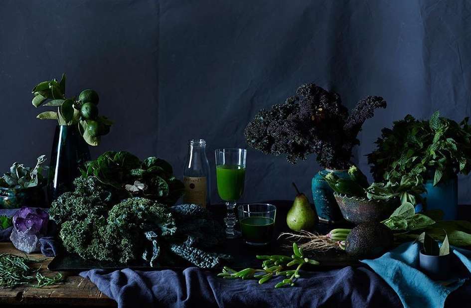 Still life photograph of green smoothies, kale, herbs, and fruit on a wooden table with a dark blue background, illuminated by natural light.