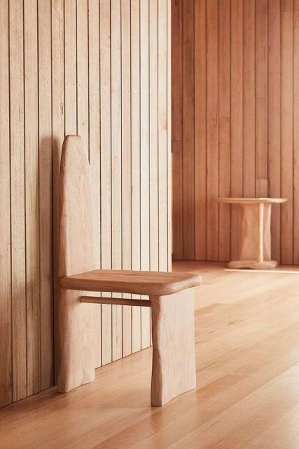 Minimalist wooden chair with a high backrest and simple table on a wooden floor in a room with light wood walls and natural light.
