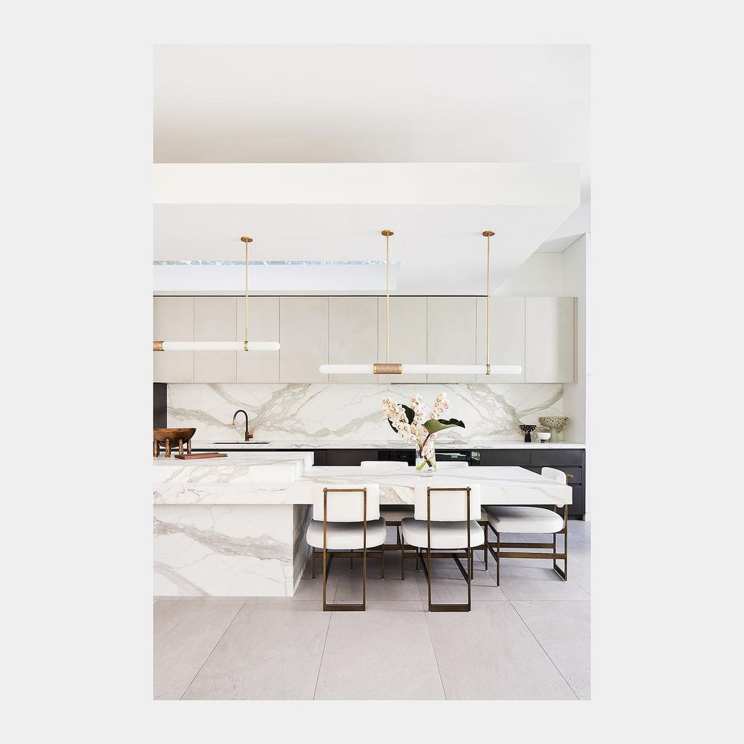 Modern kitchen and dining area featuring marble countertops, white cabinetry, gold accents, pendant lights, and seating for six.