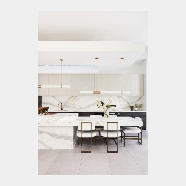 Modern kitchen and dining area featuring marble countertops, white cabinetry, gold accents, pendant lights, and seating for six.