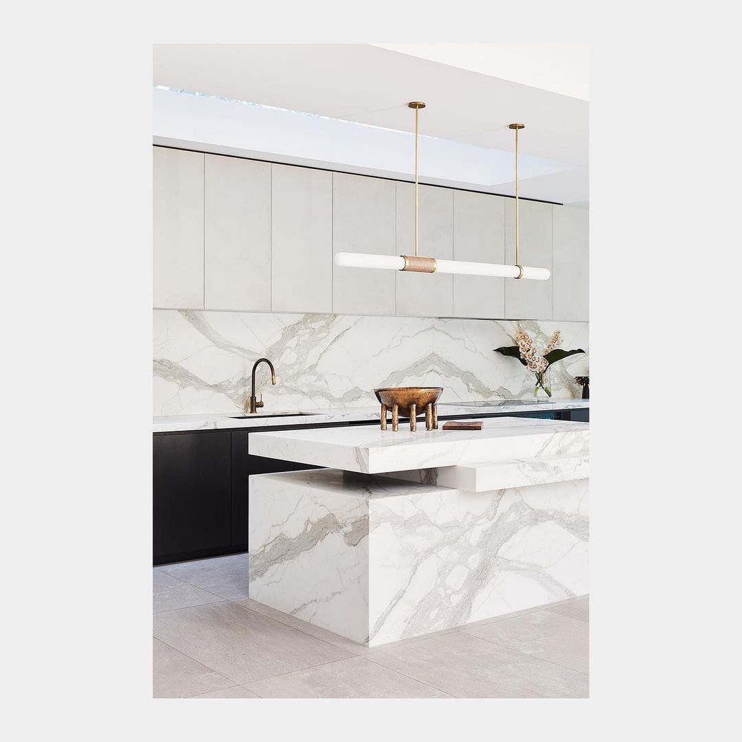 Modern kitchen interior with white marble island and countertops, stainless steel faucet, and linear lighting fixture.