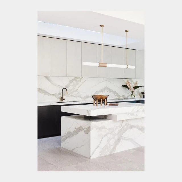 Modern kitchen interior with white marble island and countertops, stainless steel faucet, and linear lighting fixture.