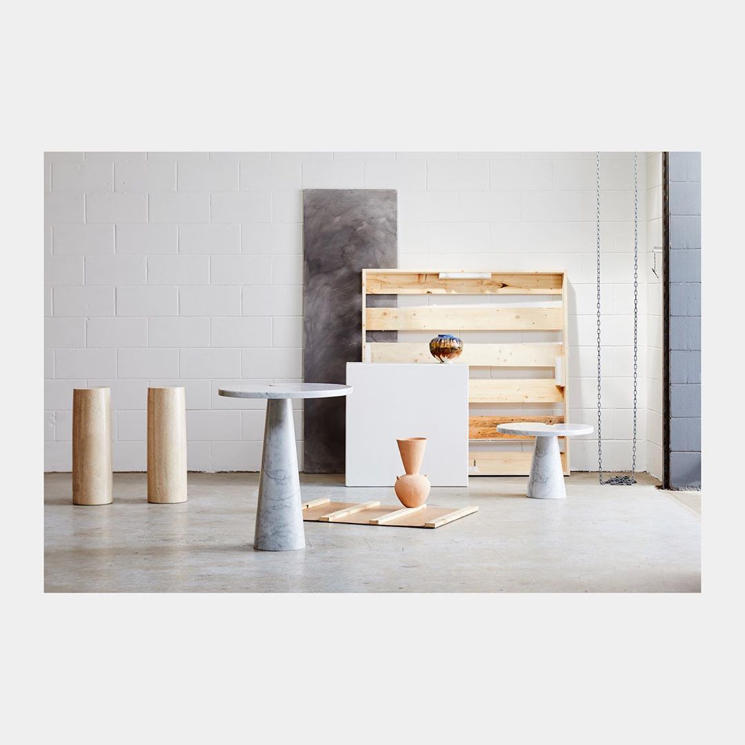 Minimalist product shot: two beige cylindrical podiums, two marble side tables, vase, and geometric shapes against white block wall.
