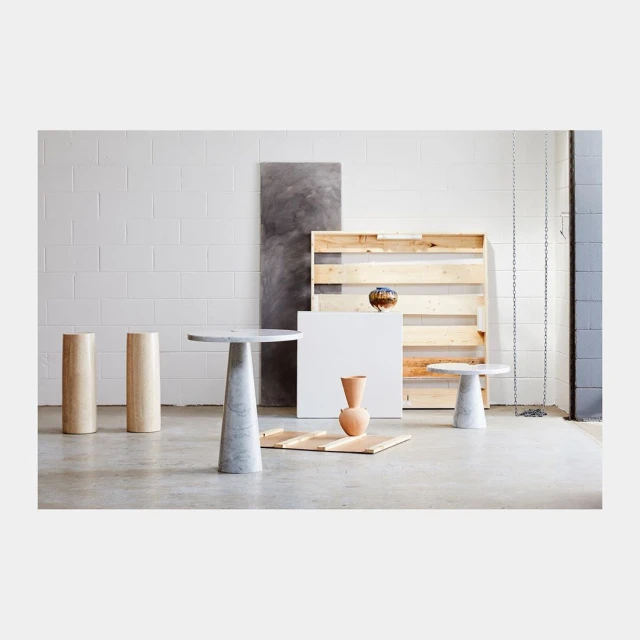 Minimalist product shot: two beige cylindrical podiums, two marble side tables, vase, and geometric shapes against white block wall.