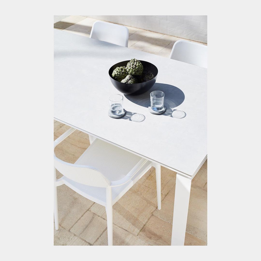 Minimalist shot of a white outdoor table with a black bowl of green plants, glasses, and white chairs on a stone tile patio.