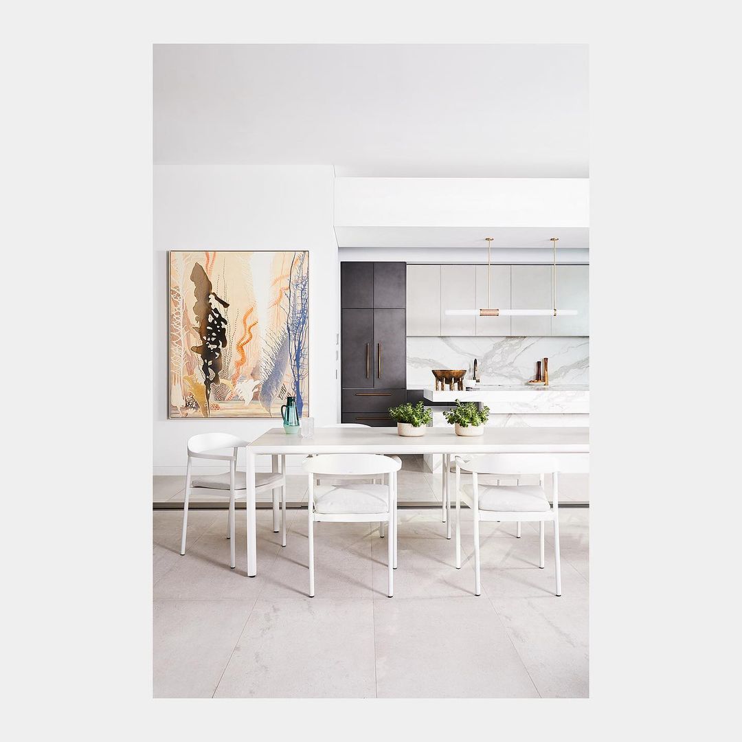 Interior dining area with white table and chairs, accented by an abstract painting, in a modern home with a kitchen background.