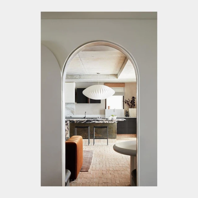 View of a modern kitchen interior with an island, pendant light, and terracotta tile flooring, framed by a white arched doorway.