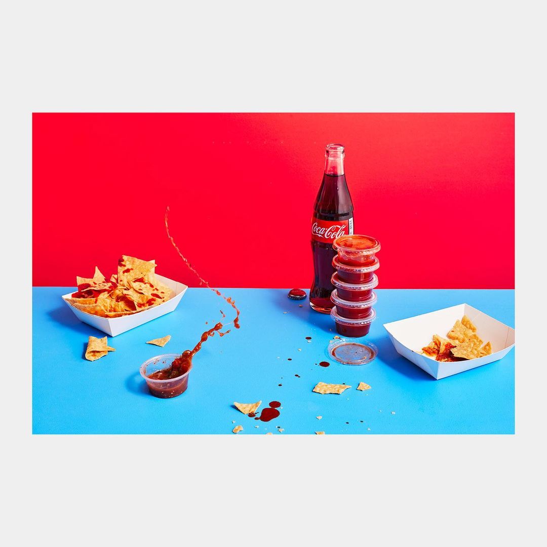 Chips and salsa served with Coca-Cola, featuring a splash of salsa with a bright red and blue background, arranged on a white surface.