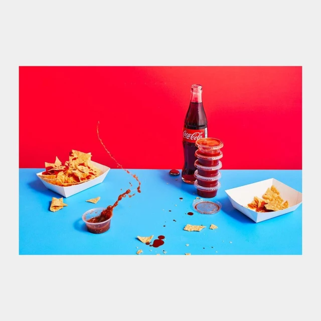 Chips and salsa served with Coca-Cola, featuring a splash of salsa with a bright red and blue background, arranged on a white surface.