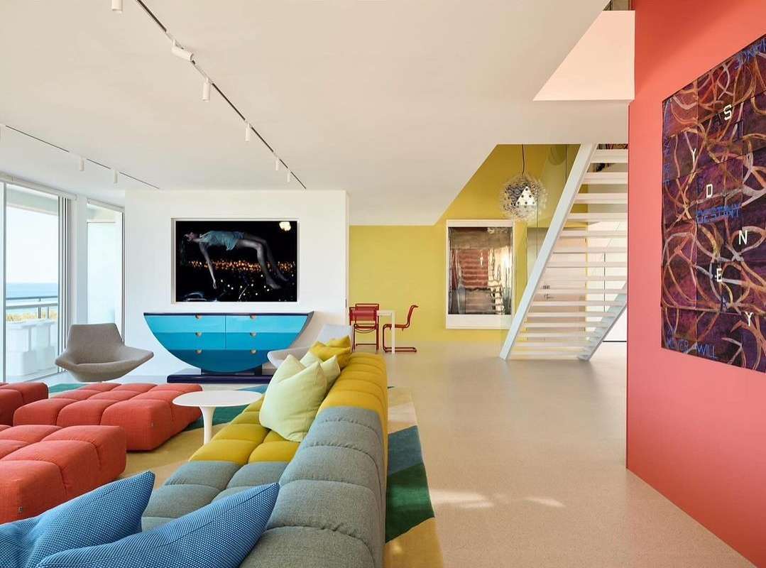 Living room interior with blue, yellow, and orange modular sofa, mid-century modern furniture, and artwork in a brightly colored space.