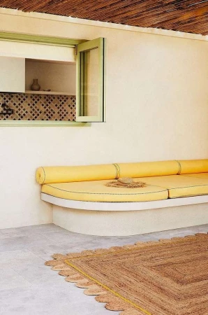 Indoor lounge area with a built-in yellow couch, natural light, a window, and a textured brown rug on a light grey floor.
