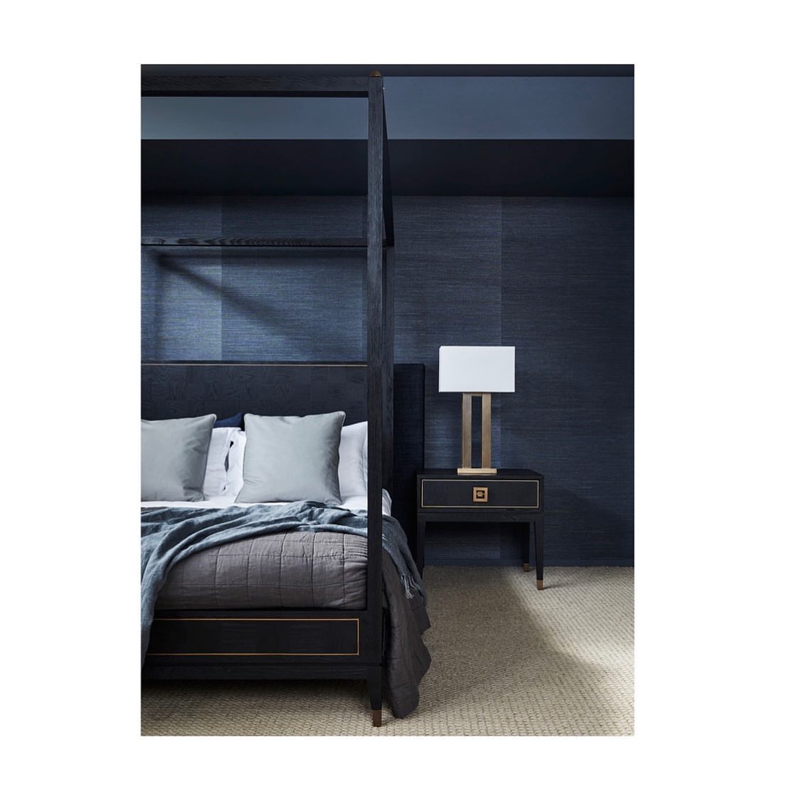 Bedroom interior with a four-poster bed, gray bedding, a bedside table with a lamp, and dark blue patterned wallpaper.